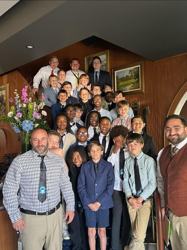Group photo of adults and children in suits and ties standing in a lobby. A flower arrangement in the corner.