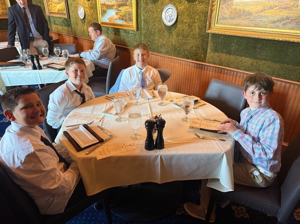 Five boys in formal attire seated at a table in a restaurant with greenery and framed art on the wall.