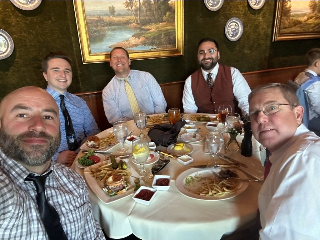 Five men are seated around a table with plates of food, glasses, and condiments. A green wall with picture frames is behind them.