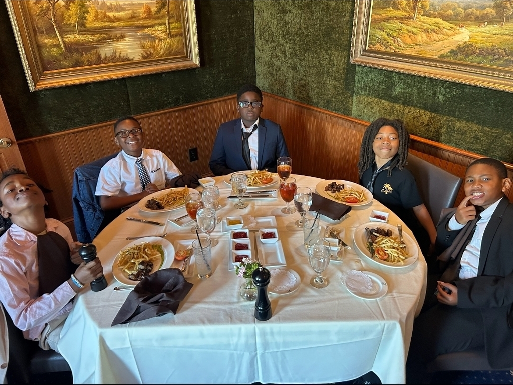 Four people sit around a table filled with food, drinks, and a bottle. A wall with two framed pictures is in the background.