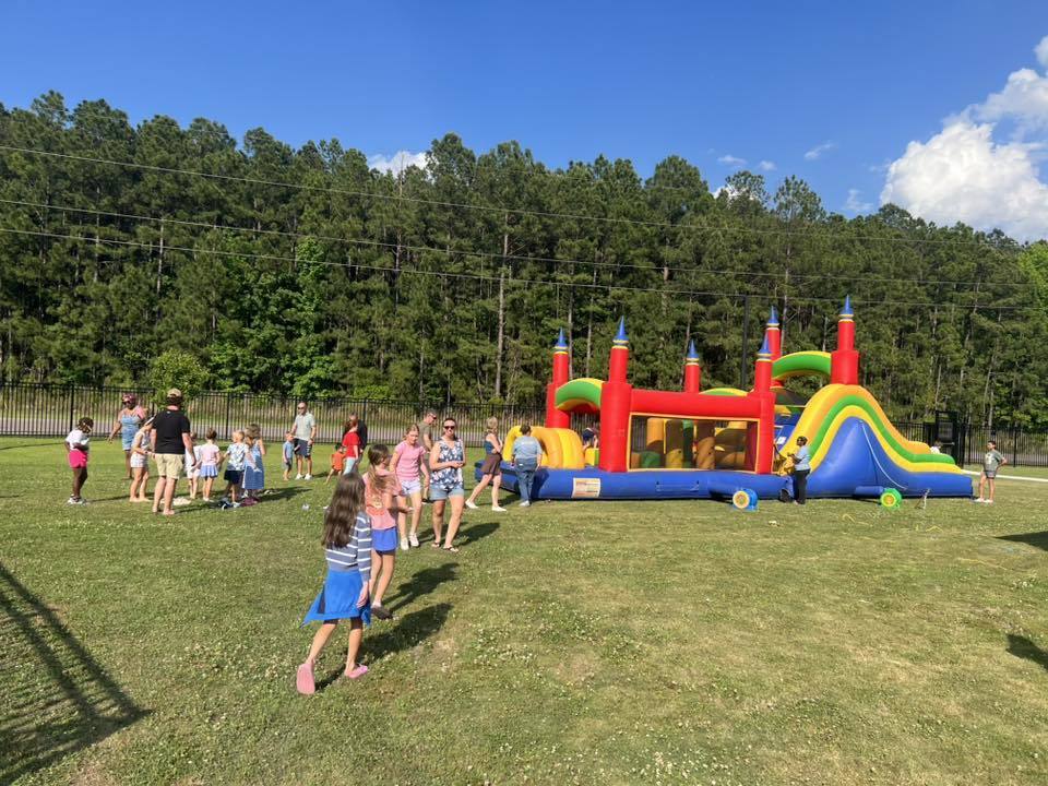 Group of people at an outdoor event, some playing near a colorful inflatable bounce house, with a grassy field and trees in the background.