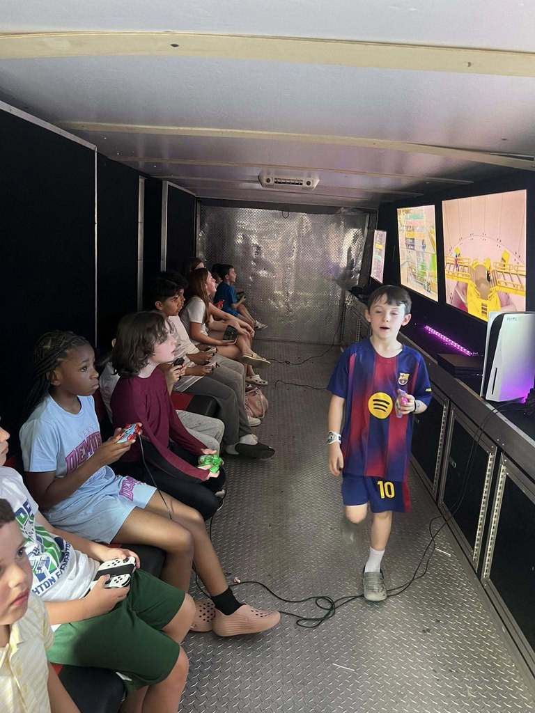 A group of kids are sitting in a mobile gaming setup, with one child wearing a soccer jersey walking towards a screen.