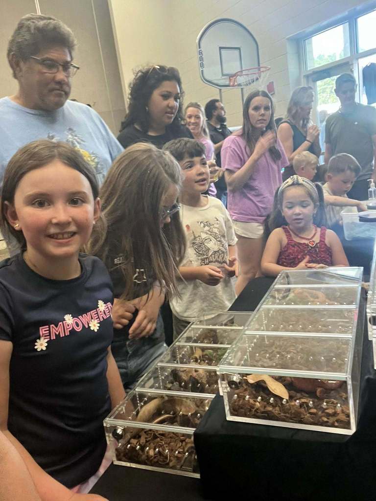 Children observe insects in glass cases at an event. Adults and other children surround them.