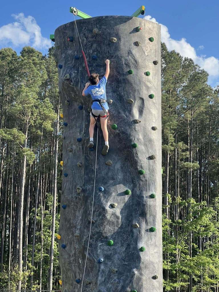 Person climbing a tall rock wall outside, secured with ropes, with a background of tall trees.