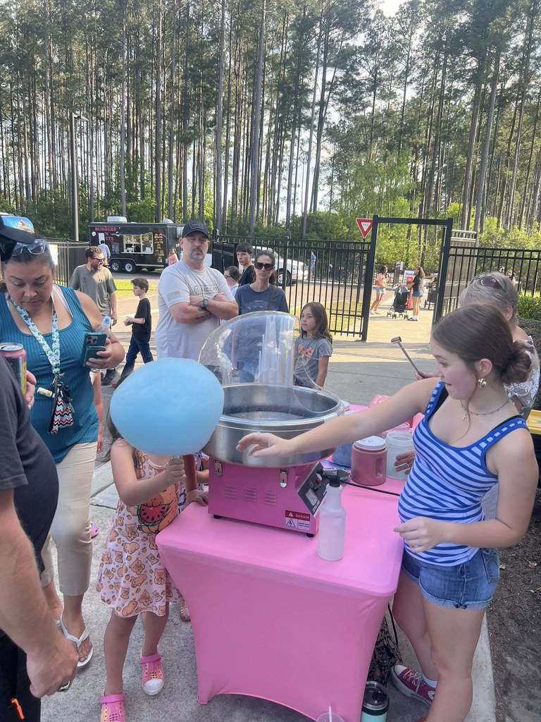 People around a pink cotton candy machine, one girl holding a blue balloon. Behind them, a fence and trees.