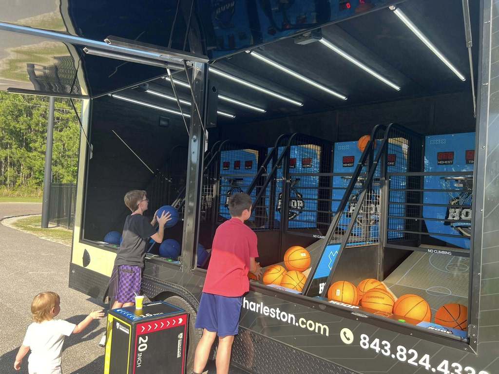 Children playing basketball on a truck-based game with multiple hoops and orange balls. The truck has a website and phone number.