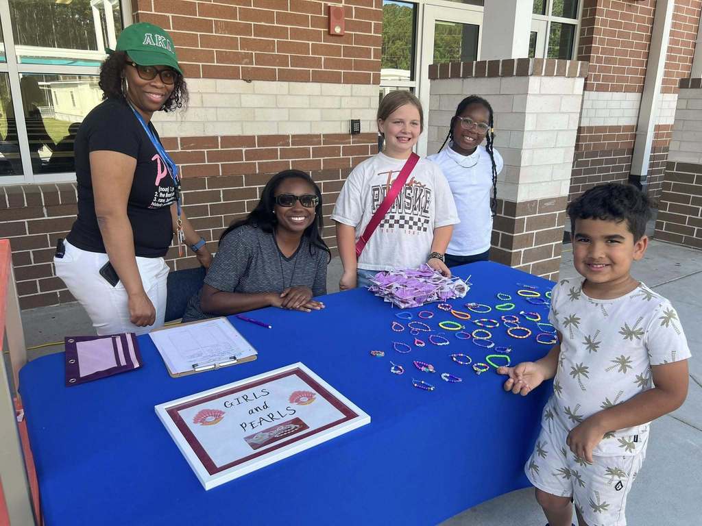 Five people gather around a table with various items, including a framed award and crafts. Two adults stand behind the table.
