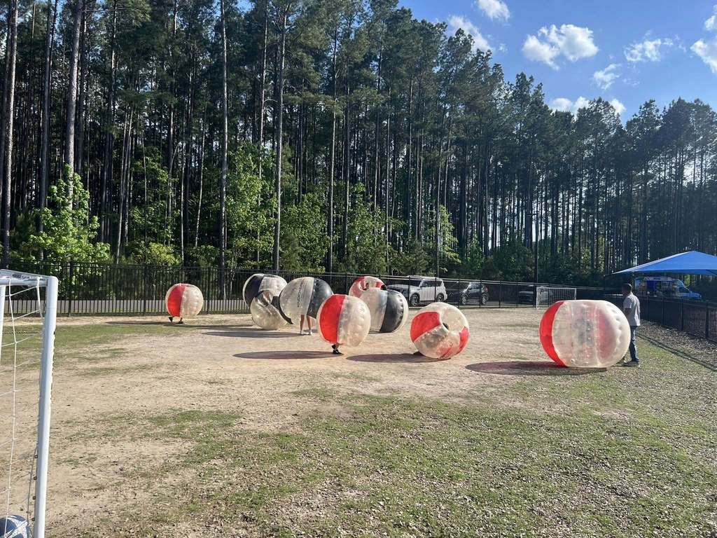 Open field with eight people in zorb balls. One person stands at a soccer goal. Trees line the perimeter.