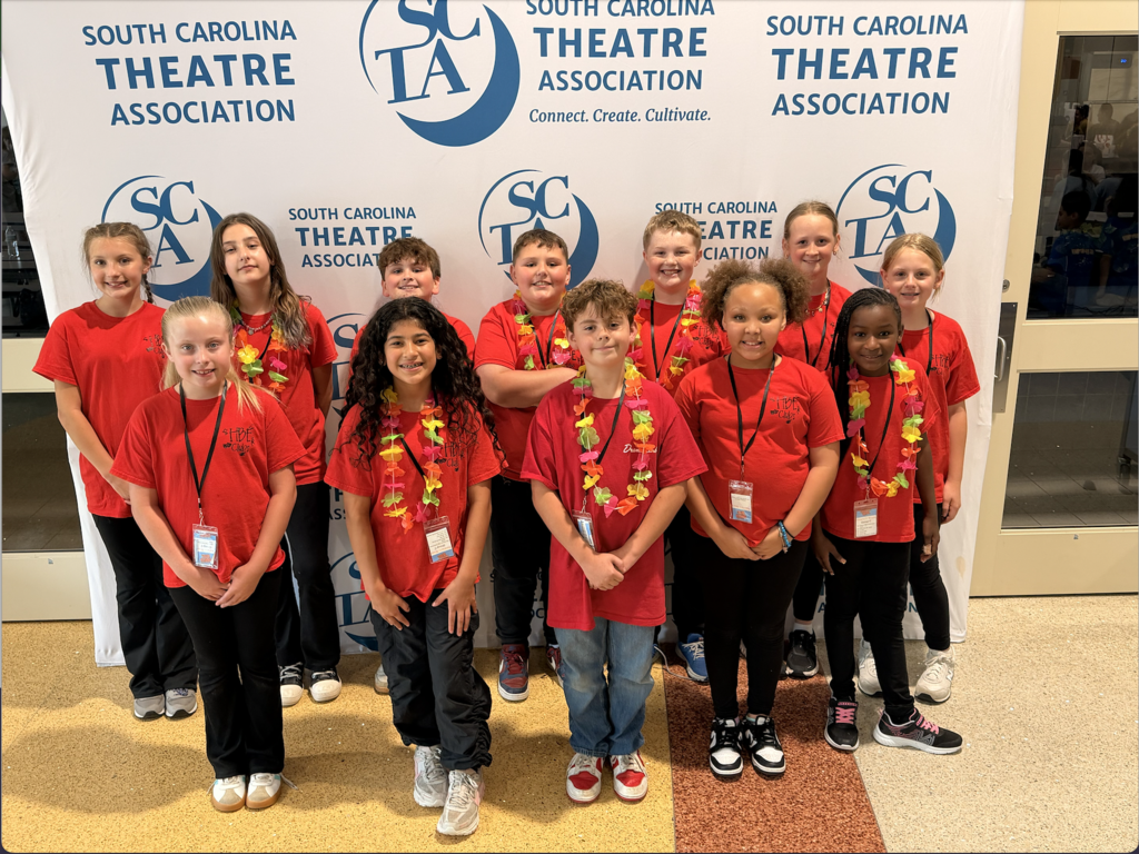 A group of approximately 12 smiling children and pre-teens wearing red t-shirts and colorful leis pose together in front of the South Carolina Theatre Association banner featuring the SCTA logo and tagline "Connect. Create. Cultivate."