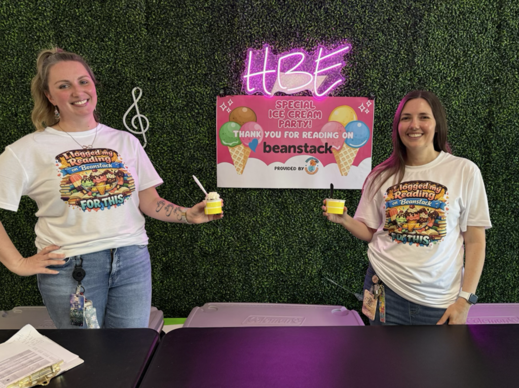 "Two smiling women stand behind a table holding small cups of ice cream. They wear matching white t-shirts with colorful graphics and text reading 'I logged my Reading on Beanstack FOR THIS.' Behind them is a green artificial hedge wall with a pink neon 'HBE' sign and a white musical note symbol. A pink banner below reads 'HBE SPECIAL ICE CREAM PARTY! THANK YOU FOR READING ON beanstack PROVIDED BY' with a small logo. Two gray Coleman coolers sit on the table in front of them."