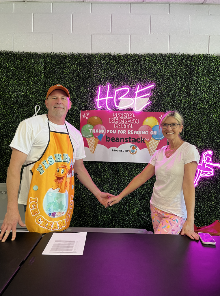 "A man and woman stand behind a black table holding hands, with a green artificial hedge wall behind them. The man wears a white t-shirt, orange cap, and orange apron featuring a cartoon fish in a fishbowl with 'FISHER ICE CREAM' text. The woman wears a white v-neck t-shirt and pink pants with a colorful sprinkle pattern. A pink neon 'HBE' sign and banner reading 'SPECIAL ICE CREAM PARTY! THANK YOU FOR READING ON beanstack PROVIDED BY' are visible on the wall, along with a partial neon ballet dancer sign. A document and smartphone rest on the table."