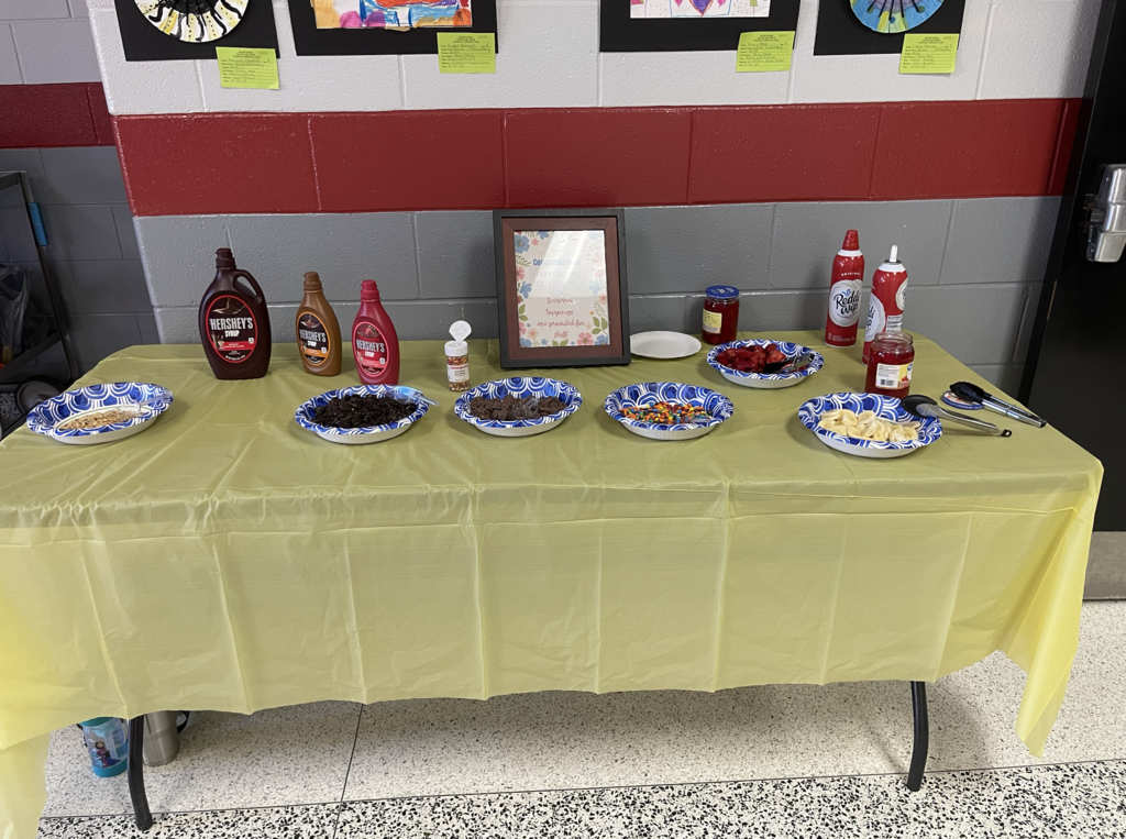 "A yellow tablecloth-covered table displays an ice-cream topping bar against a red and gray wall. The table features three Hershey's syrup bottles (chocolate, caramel, and strawberry), two Reddi-wip whipped cream cans, and paper bowls containing chopped nuts, crushed cookies, chocolate chips, sprinkles, multi-colored candies, sliced strawberries, and sliced bananas. Two jars of red fruit preserves and serving utensils (tongs and spoon) are also visible. A framed sign reads 'Choose to be happy' and 'Always be grateful for small things.' Children's artwork hangs on the wall above the table with yellow sticky notes. A blue 'Frozen' character cup sits on the gray and white tiled floor below the table."
