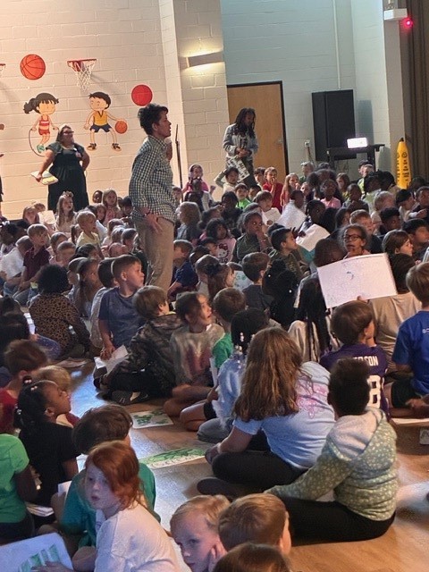 A large group of children sit on the floor of a room with adults. Some children hold signs.