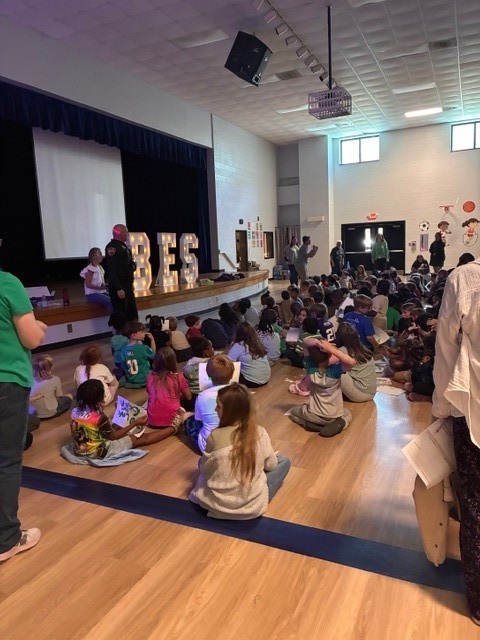 Group of children sitting on floor in room with stage, spotlights, and a projector screen.