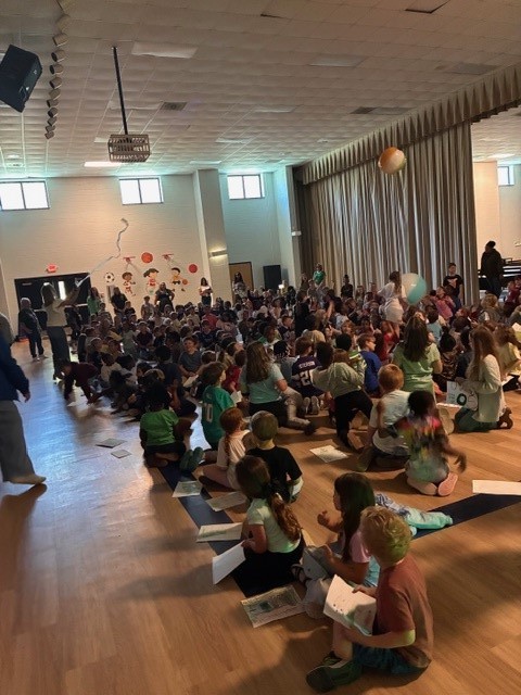A large group of people, some seated on the floor and others standing, in a room with wooden floors.