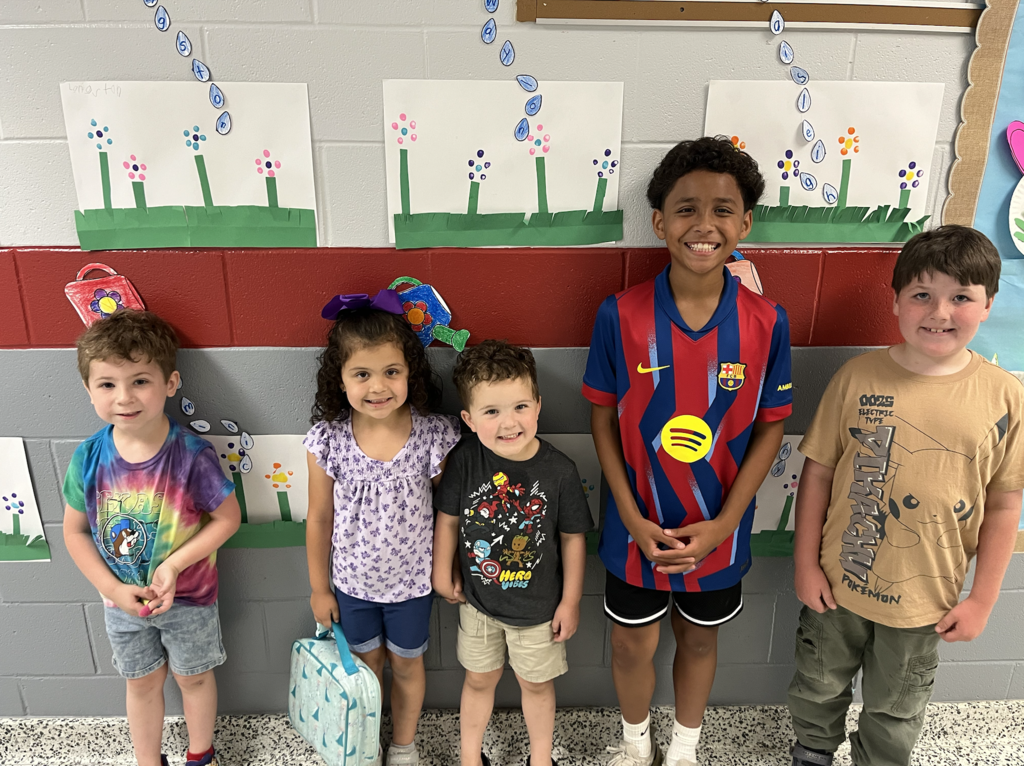 Five children, smiling and standing together in front of a wall decorated with flower paintings.