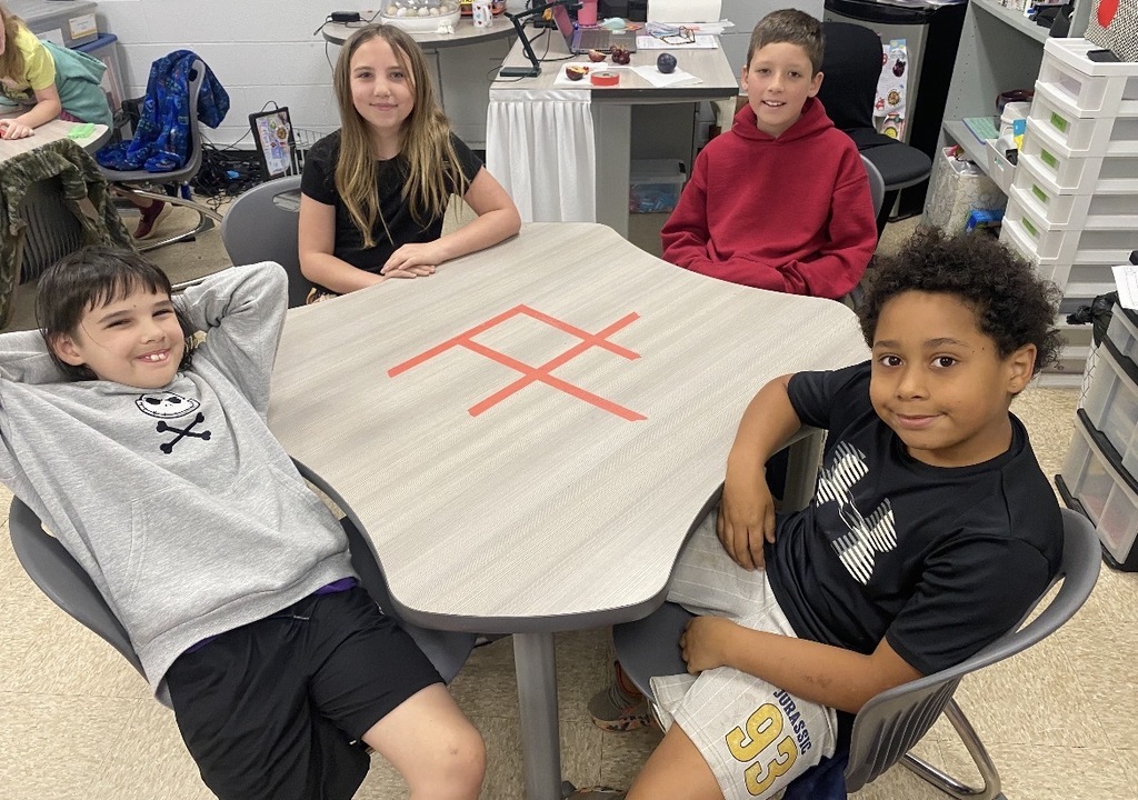 Four children sit around a table, posing for a photo. A red symbol is on the table's surface.