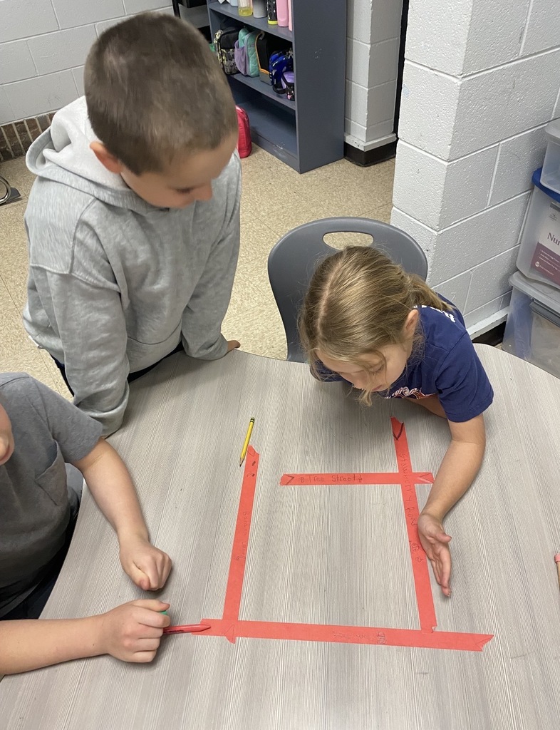 Three children at a table, using colored tape to create a shape. A boy and a girl are standing, another sits.