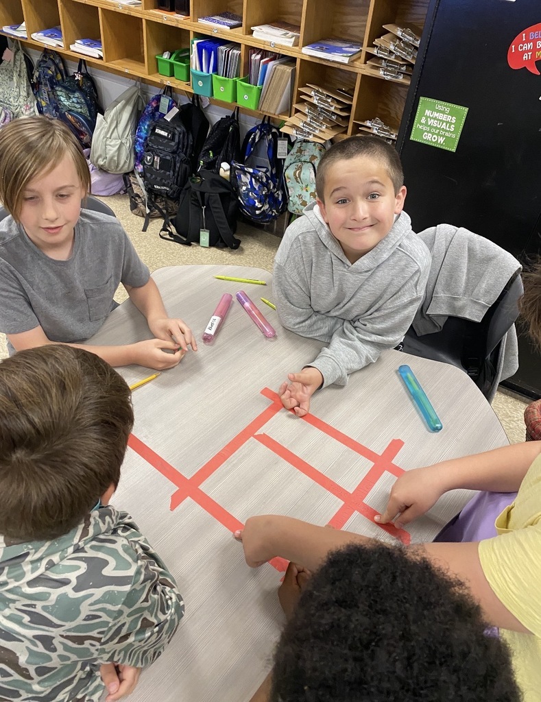 Four children in a classroom sit around a table, drawing with colored markers on a red grid.