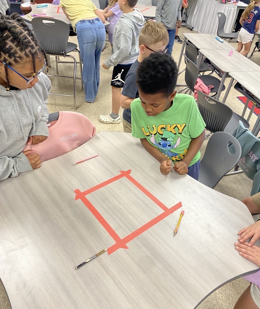Children work at a table in a classroom. One child wears a green shirt with the word "LUCKY."
