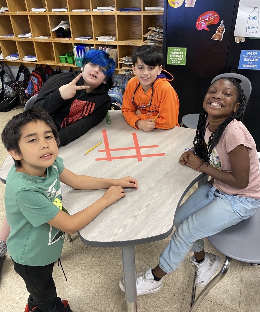 Four kids sit at a table in a classroom. One has blue hair, one wears orange, and one wears a pink shirt.
