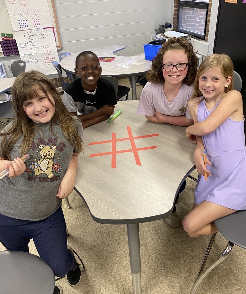 Four children sitting at a table, one drawing a hashtag on it. The room has chairs, tables, and a whiteboard.