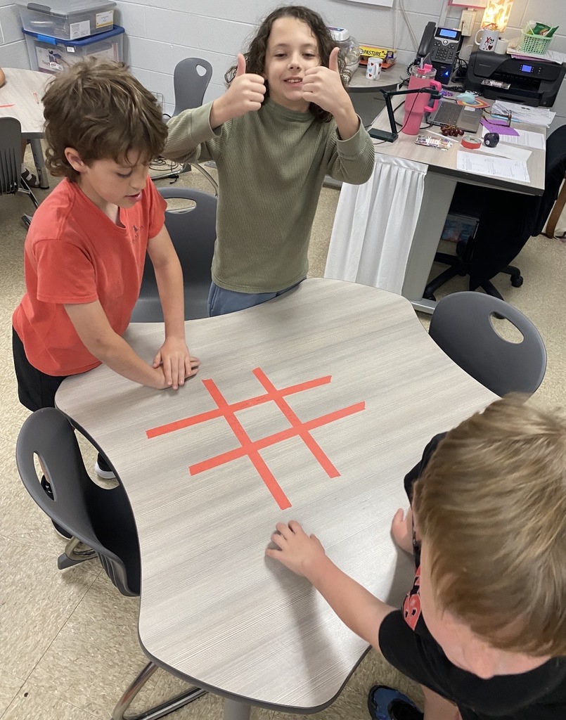 Three children in a room, playing a game on a table with a red hash symbol.