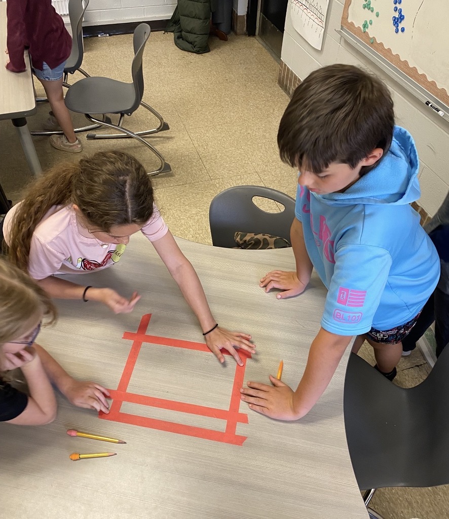 A classroom scene where three children are seated at a table. One child draws on a red paper square with markers.