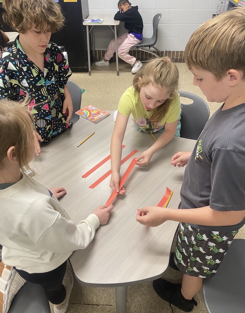Four children are seated at a table, working together on a project involving red paper.