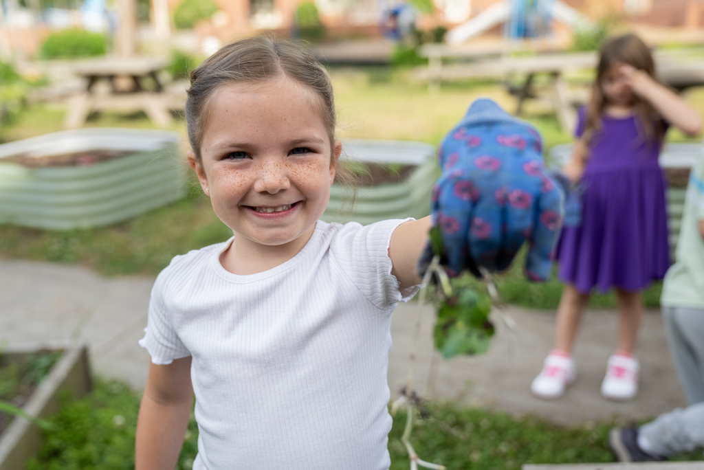 A young girl with gloves holds a plant. Behind her, two others look at something. Raised garden beds and a playground are in the background.