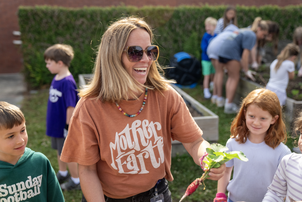 A woman wearing a shirt with "Mother Earth" text holds a radish. She stands with three children, two of whom wear bracelets.