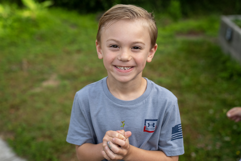 Boy holds plant, smiling, with teeth showing, on green lawn. 