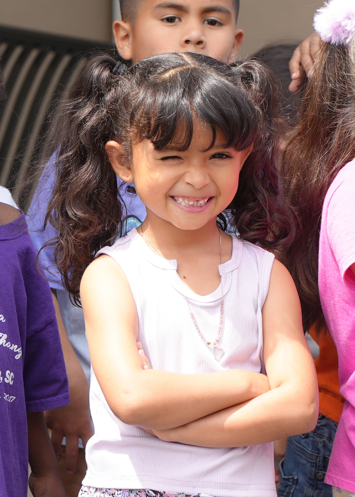 Girl with short black hair in a white sleeveless top stands with arms crossed and smiling. Behind her, a blurred group of children, one with a ponytail and a purple shirt.