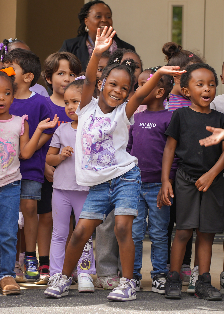 A group of children stand on pavement, one girl with her arms raised. Various children wear different colored shirts and sneakers.