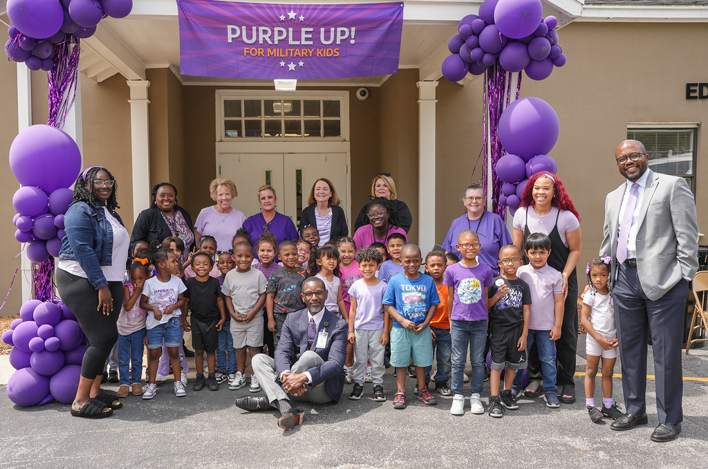 A group of adults and children pose for a photo in front of a building with purple balloons.