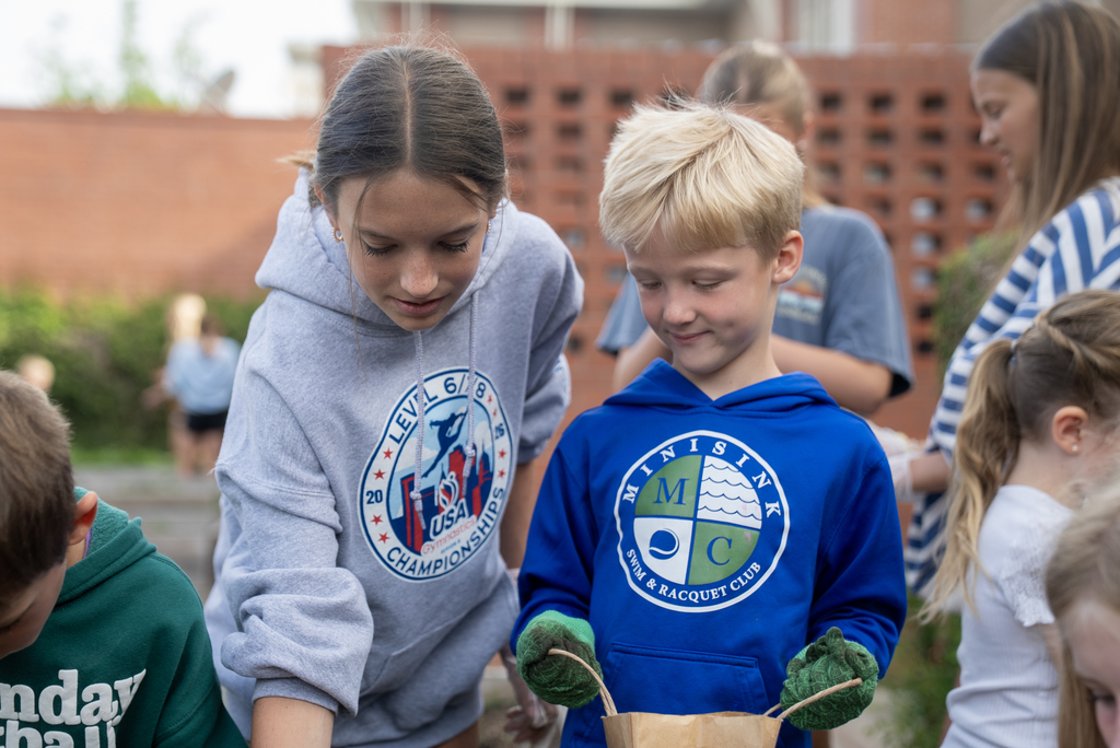 Children in hoodies work in a garden. One wears a blue hoodie with an emblem. A brick wall is in the background.