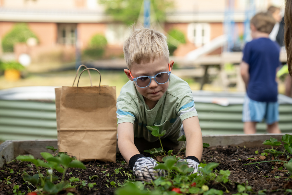 A child with glasses and gloves planting in a garden bed with a brown bag next to him.