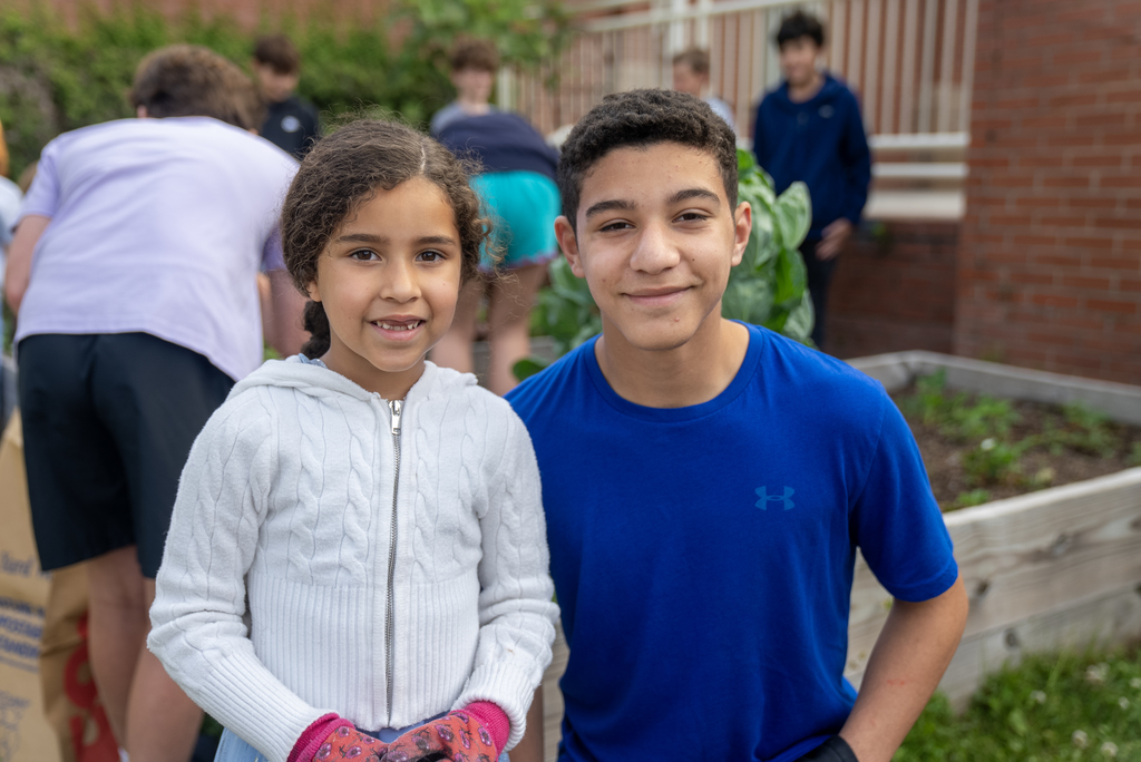 Two children, a girl in a white jacket and a boy in blue, smile for a photo in a garden.