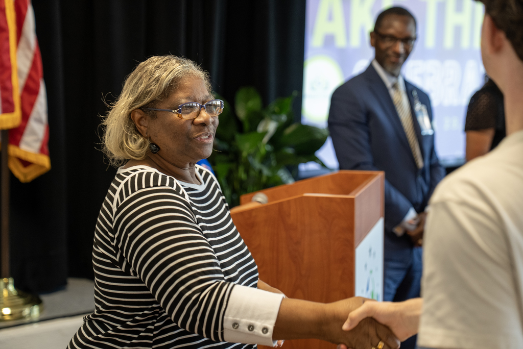 Woman in glasses and striped top shakes hands with another person, a man in a suit stands behind her.