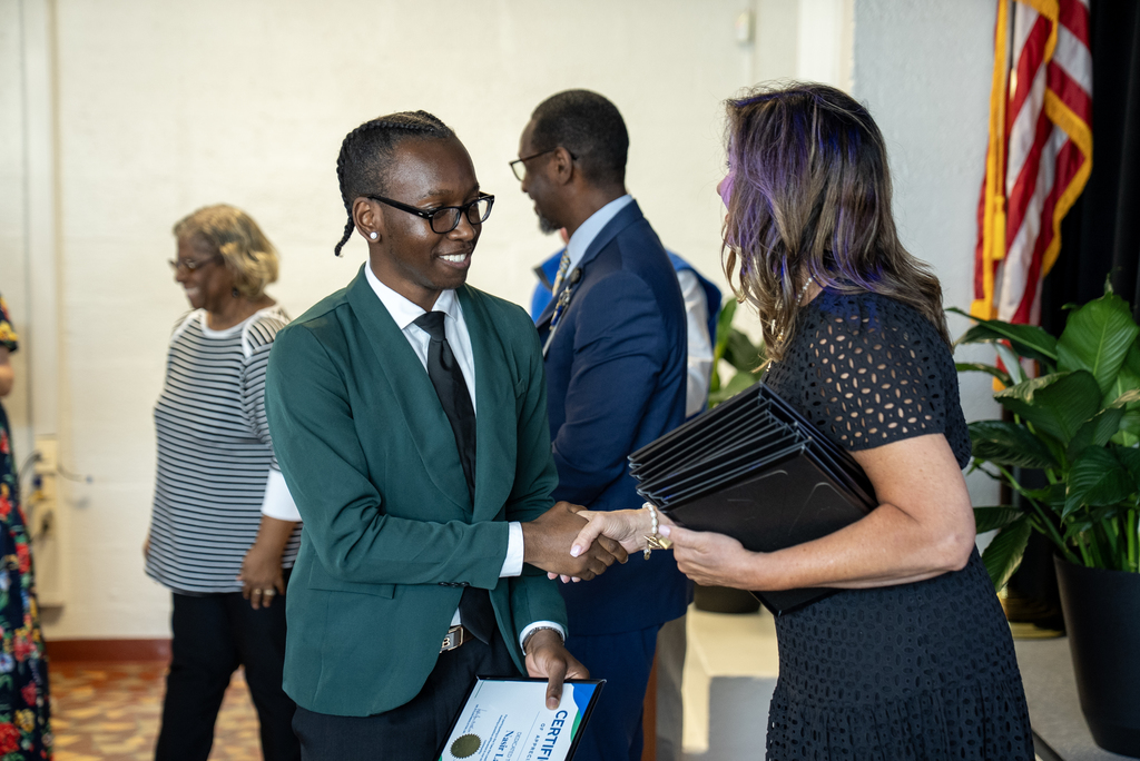 A man in a green suit and a woman in a black dress shake hands. Behind them, people stand near a potted plant.