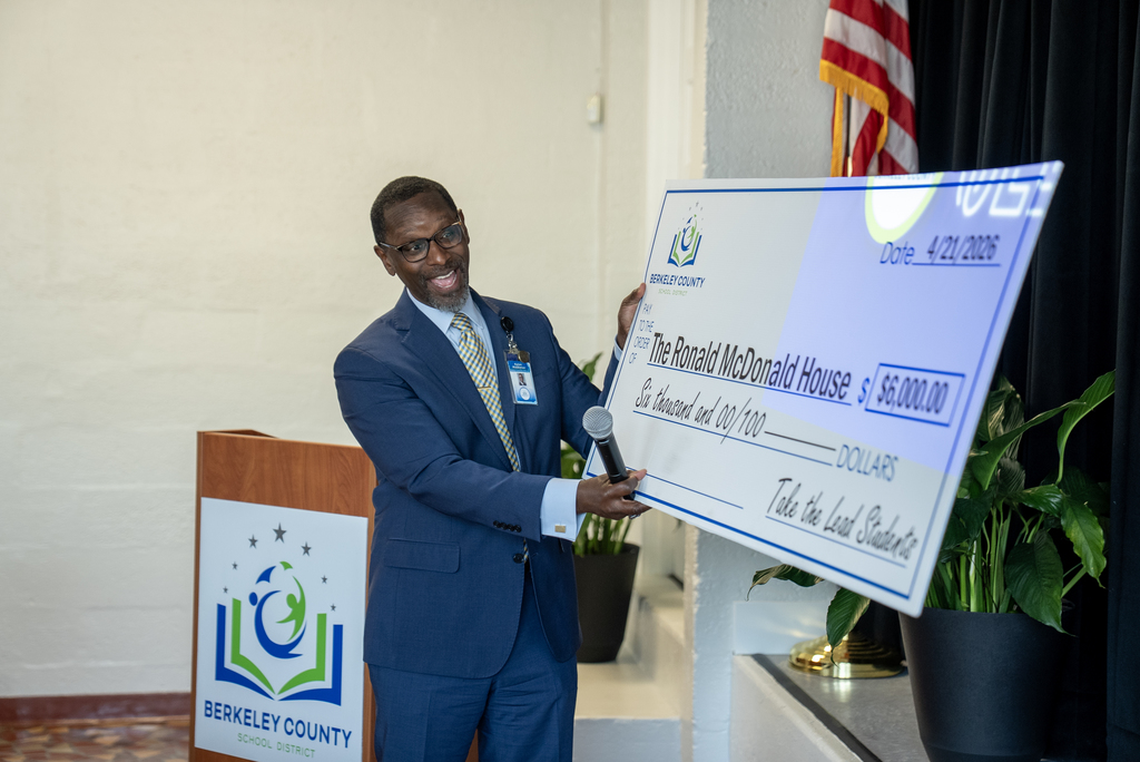 A man holding a check in a room with a BCSD logo on a podium, plants, and an American flag.