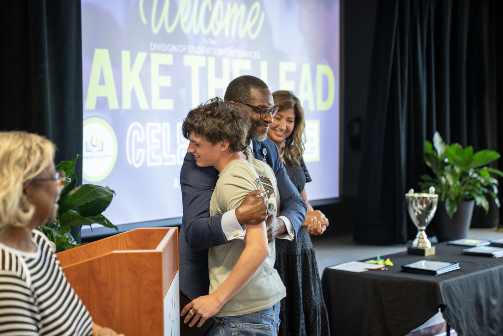 A group of people stand in a room, with one man embracing another in front of a projection screen.