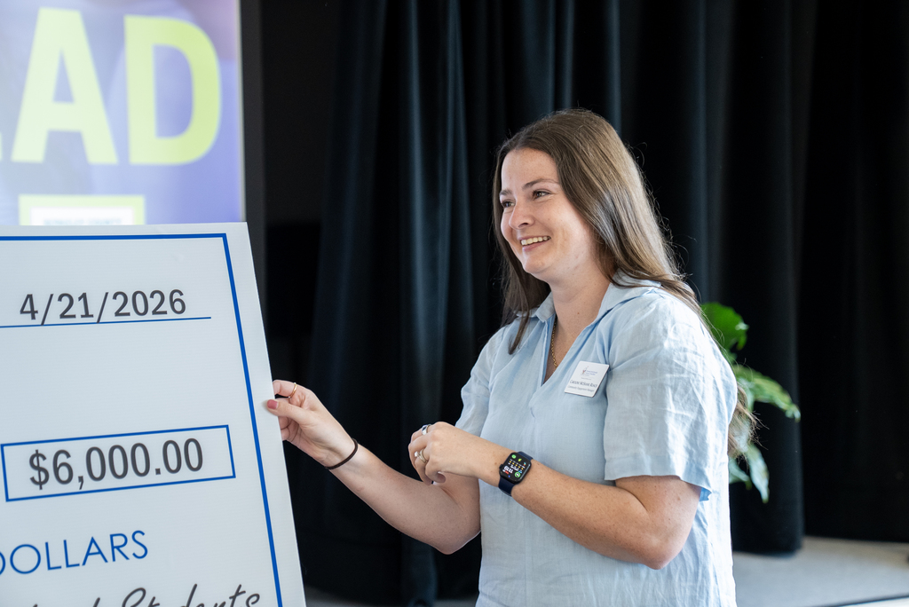A woman holds a check reading "$6,000.00" with the date "4/21/2026" on a white background. She smiles and wears a light blue shirt.