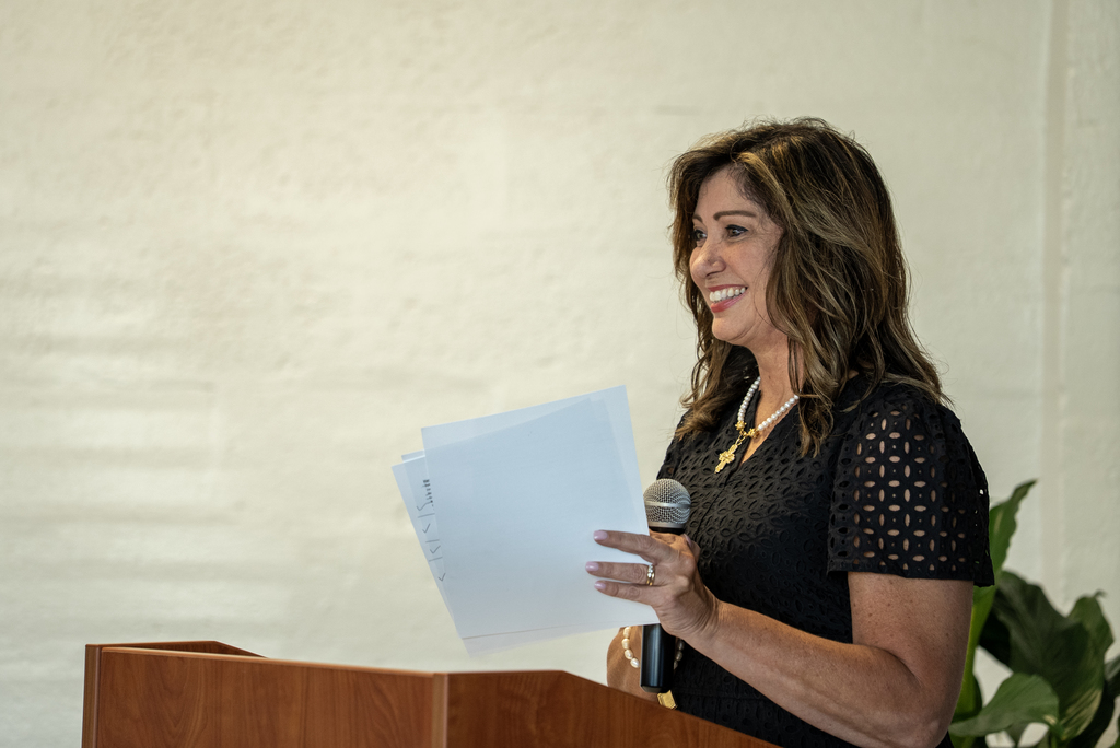 Woman at a podium, holding a microphone and papers, standing in front of a white wall.