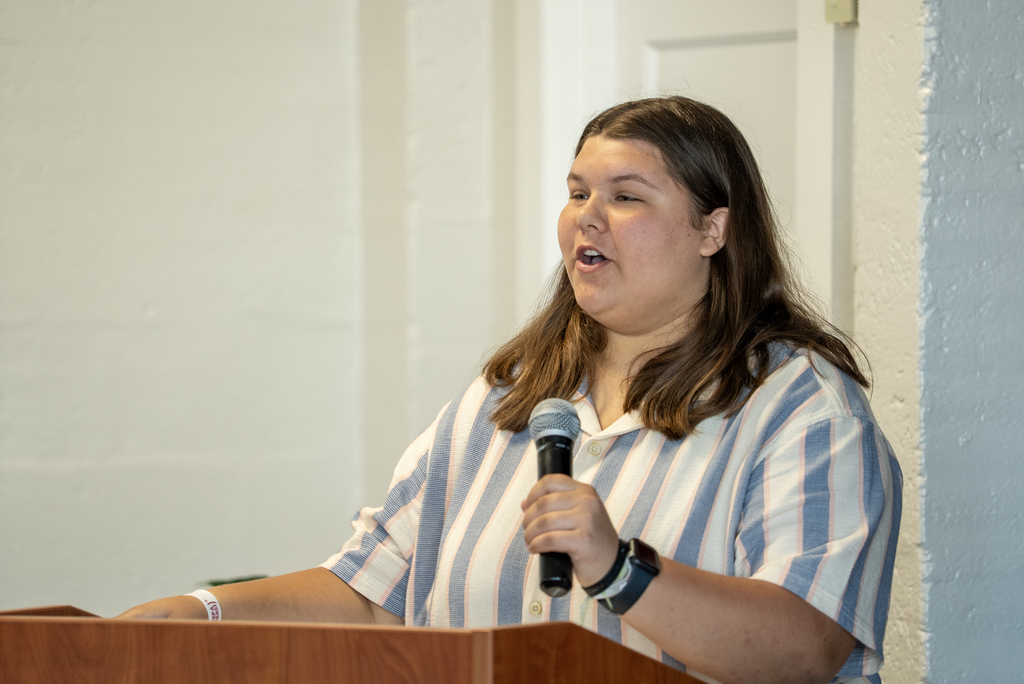 A person at a podium holds a microphone and speaks. A white wall and door are behind the person.