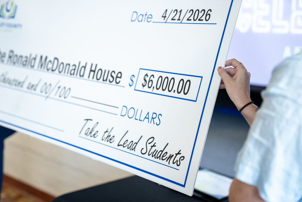 A person holds a large check for $6,000.00 made out to Ronald McDonald House, dated April 21, 2026.