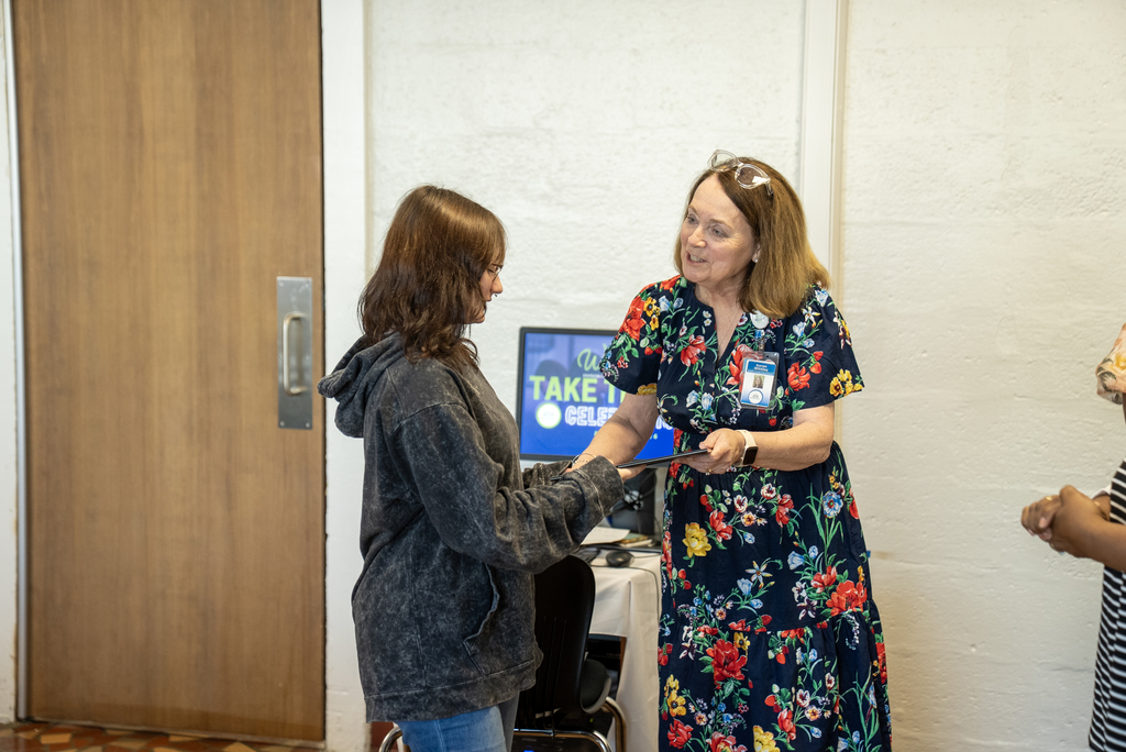 Two women in a room, one hands a card to the other.