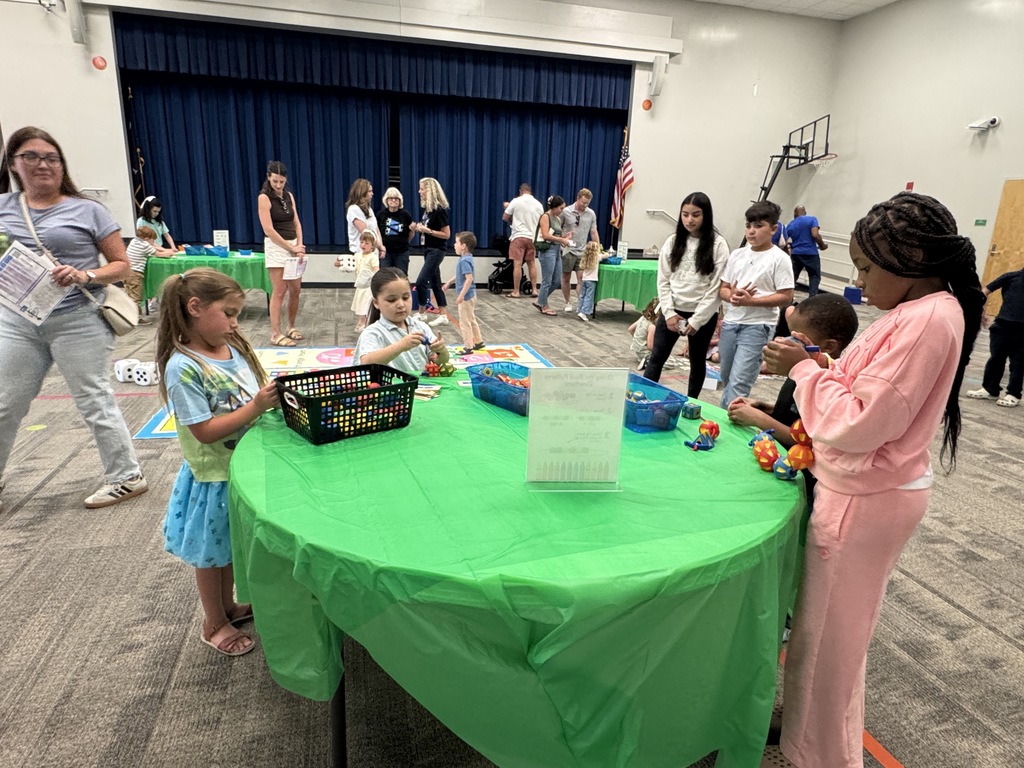 A group of adults and children in a room with a blue curtain. A woman and a girl stand at a green table.