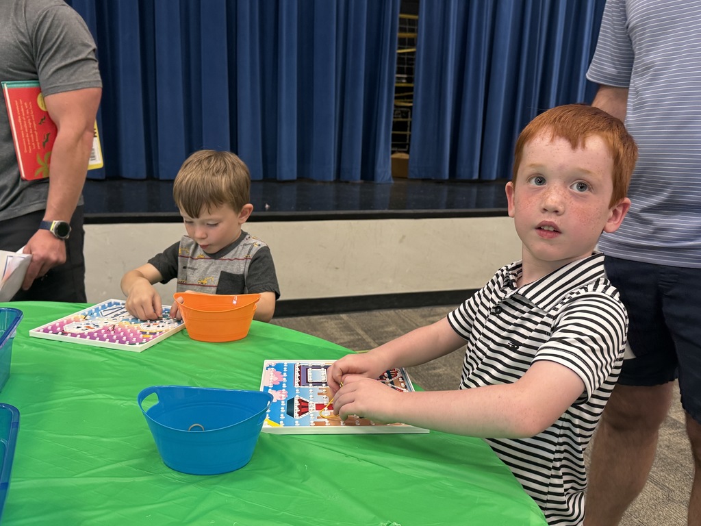Two boys playing a game at a table with an orange bowl, green tablecloth, blue bowl, and blue curtain.