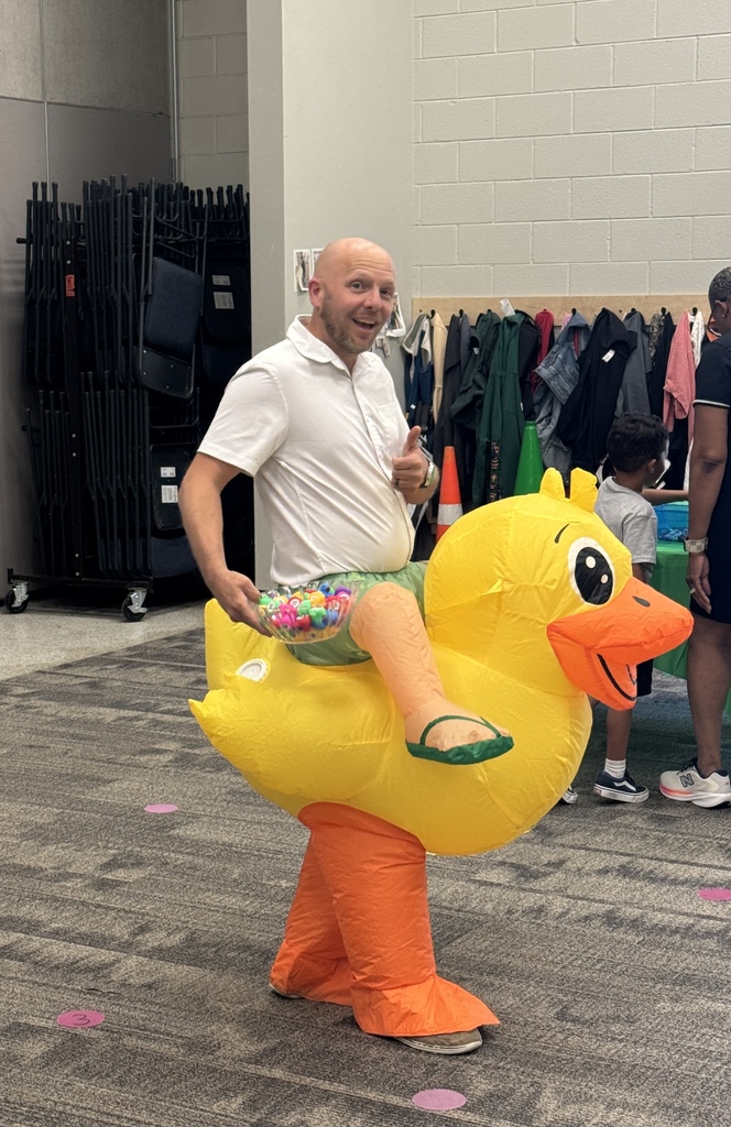 Man in white shirt and green shorts riding a yellow inflatable duck in a room with storage boxes.